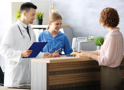 Eine Frau steht an einem Tresen zur Anmeldung und wird von einer Krankenpflegerin und einem Arzt empfangen.